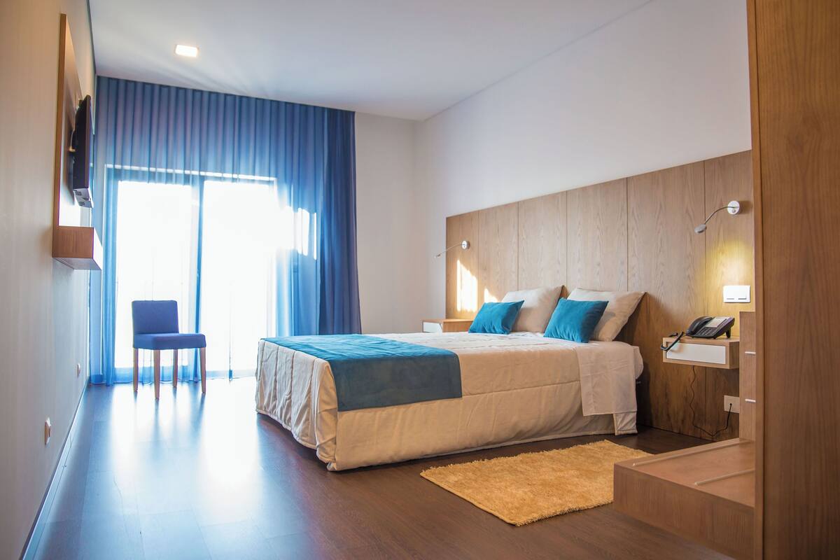A modern hotel room with a large bed, a chair, a desk with a phone, and a window with curtains.