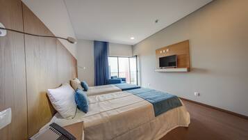 A modern hotel room with two beds, a desk with a computer, a TV mounted on the wall, and a large window with curtains.