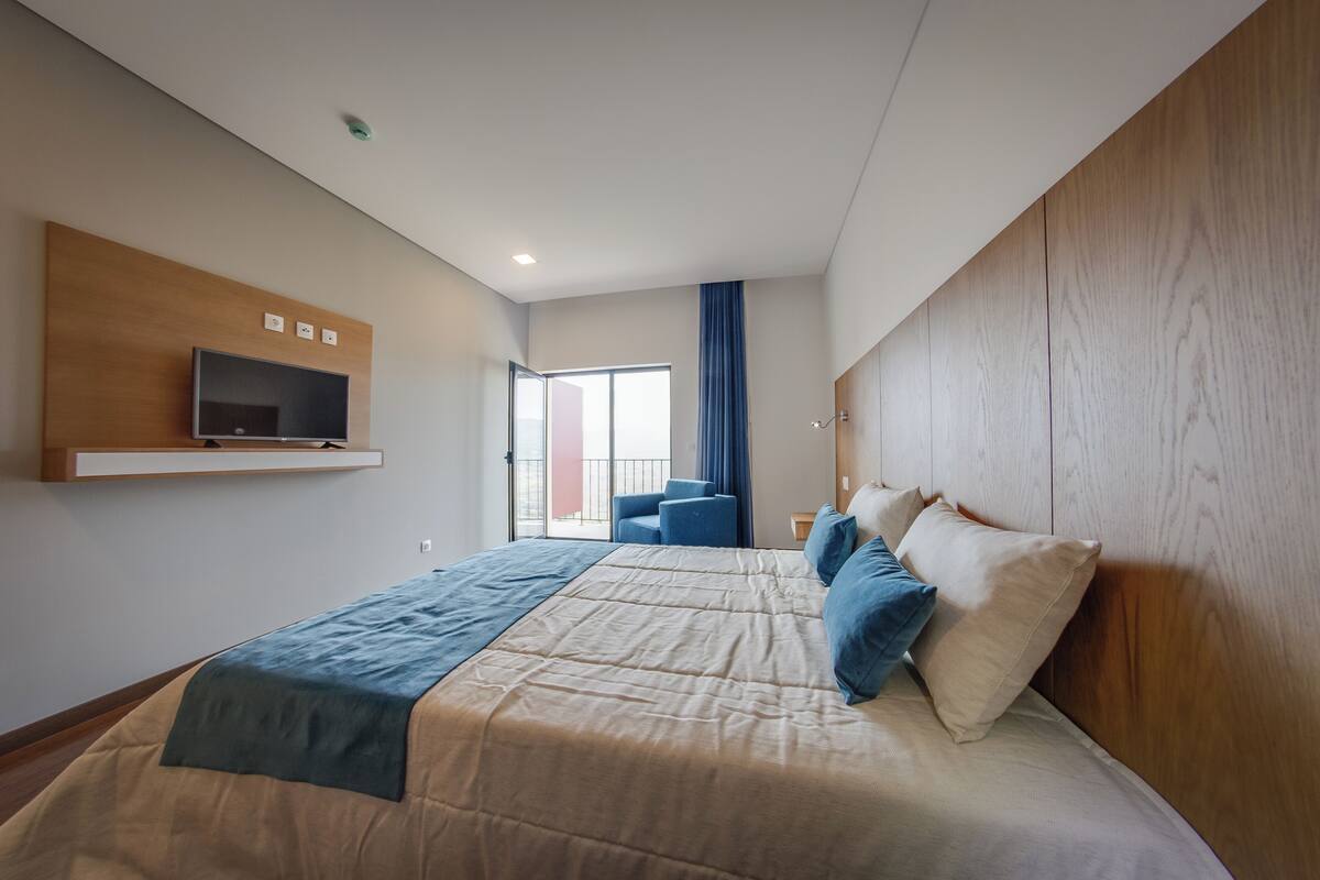 A modern hotel room with a large bed, a mounted TV, a balcony with a chair, and a wooden headboard.