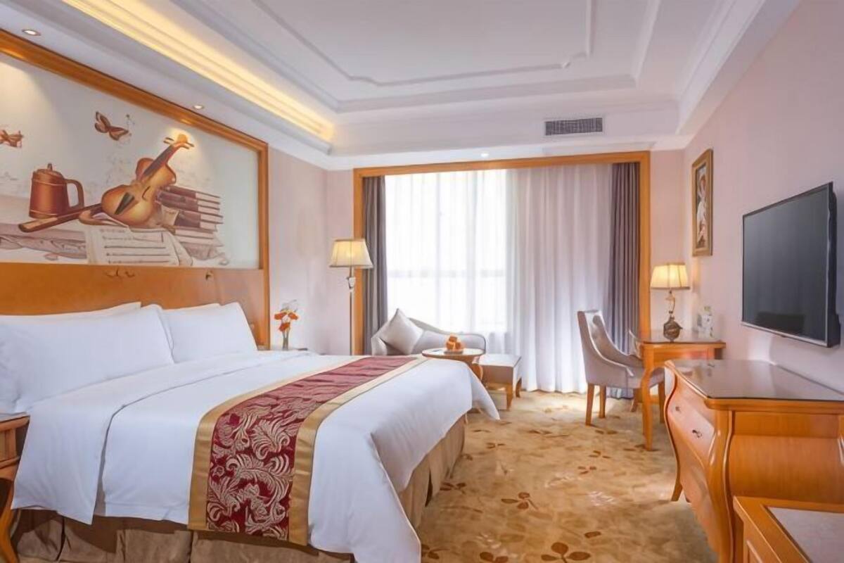 A hotel room with a large bed, a desk, a chair, a TV, and a painting on the wall.