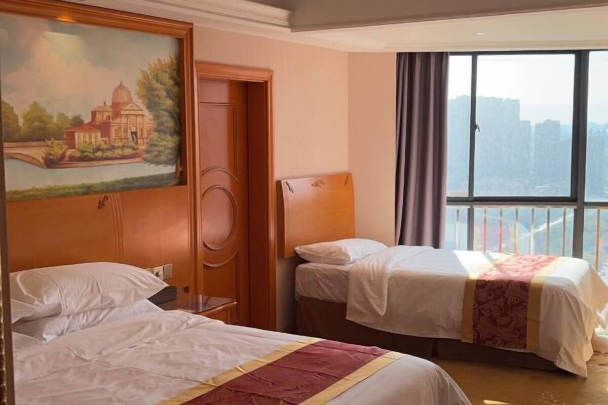 A hotel room with two beds, a painting on the wall, a window with a city view, and a door.