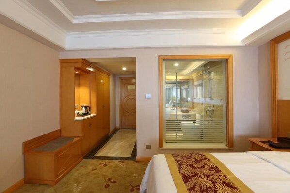 A hotel room with a bed, a bedside table, a wardrobe, a mirror, and a bathroom visible through a glass door.