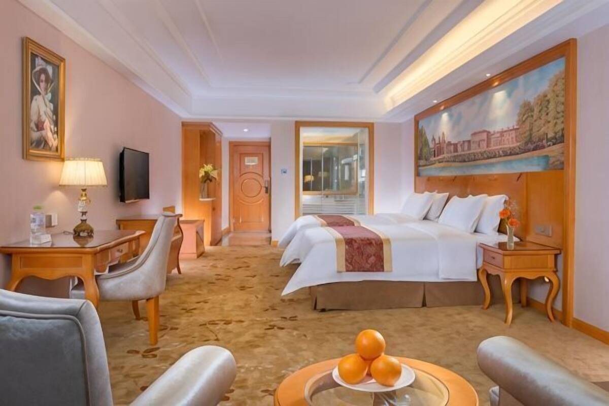 A hotel room with a bed, two bedside tables, a desk, a chair, a television, and a painting on the wall.