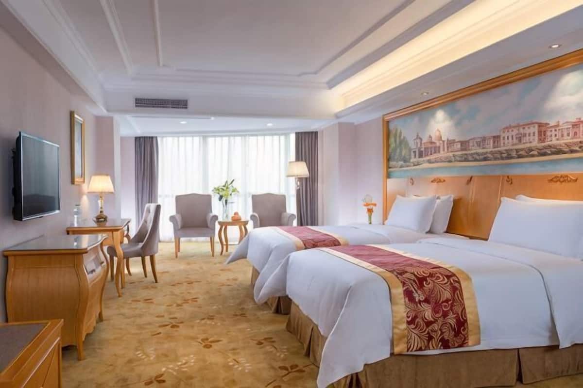 A hotel room with two beds, a desk, a chair, a TV, and a painting on the wall.