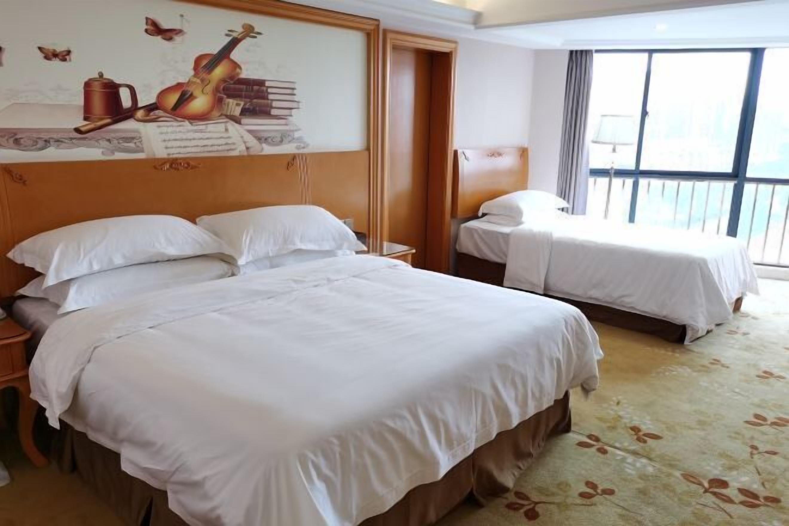 A hotel room with two beds, a large window with a view, and a wall with a musical instrument and books.