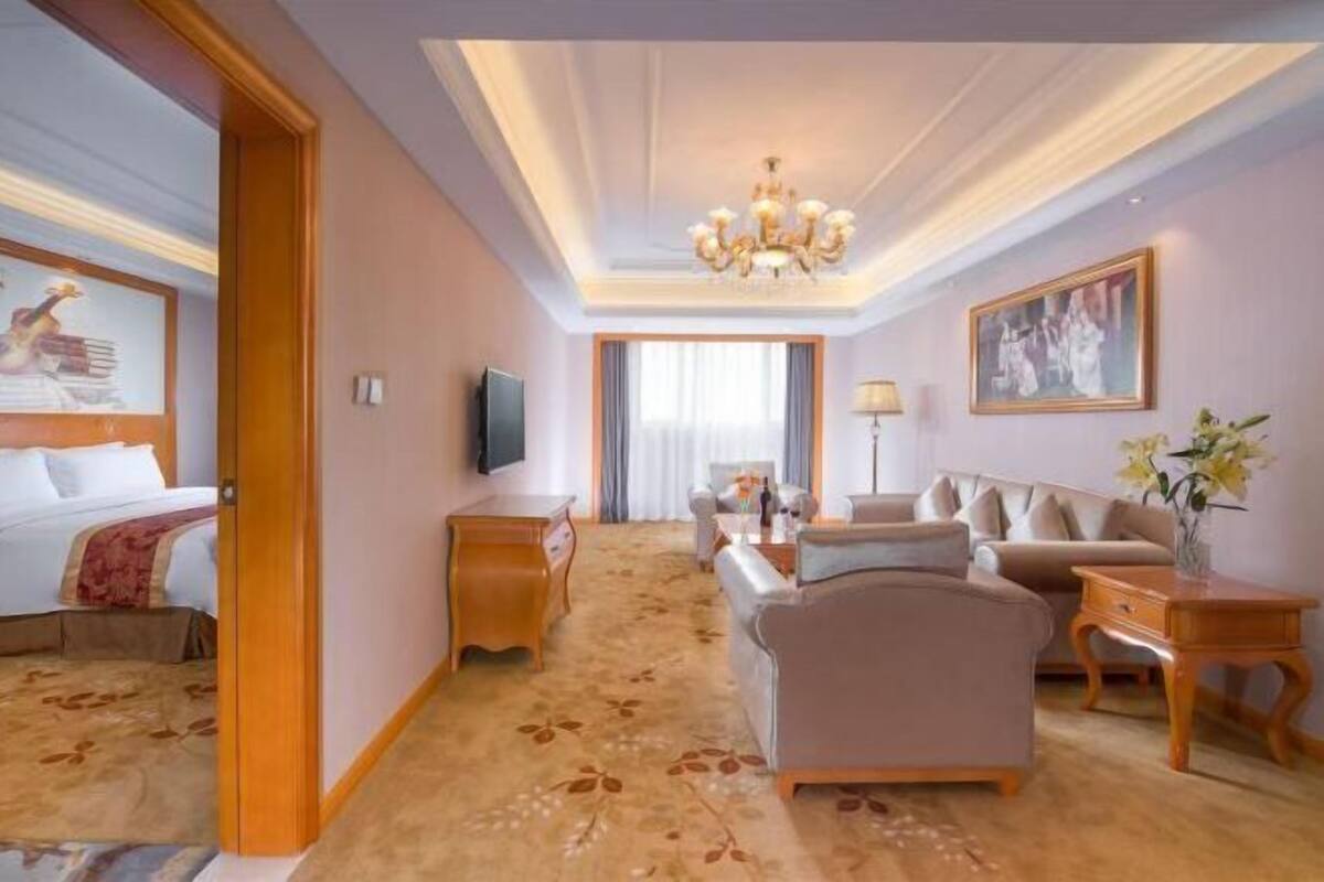 A hotel room with a bed, a sofa, a TV, a chandelier, and a painting on the wall.
