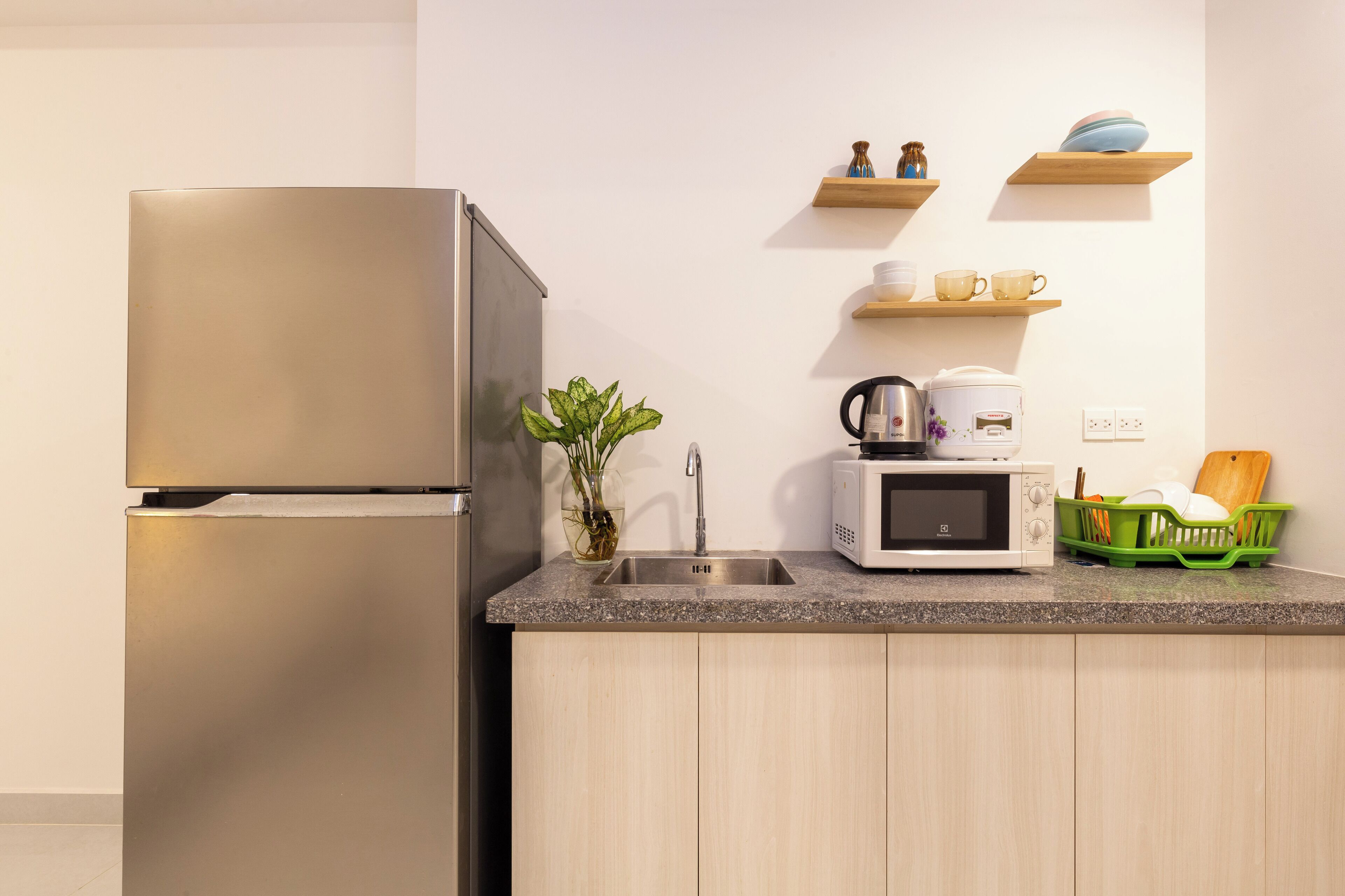 Una cocina moderna con un refrigerador de acero inoxidable, un microondas y un lavamanos. Hay repisas de madera sobre la encimera con tazas y cuencos.
