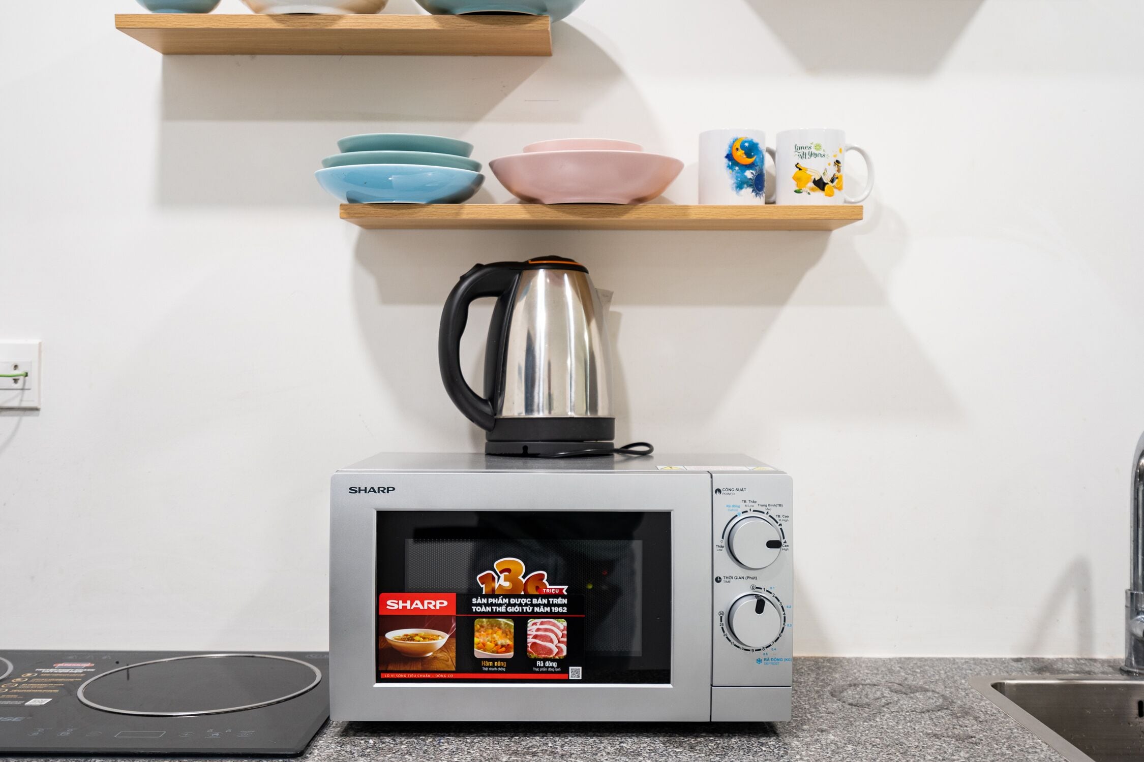 A kitchen with a microwave, a kettle, and a shelf with bowls and mugs.