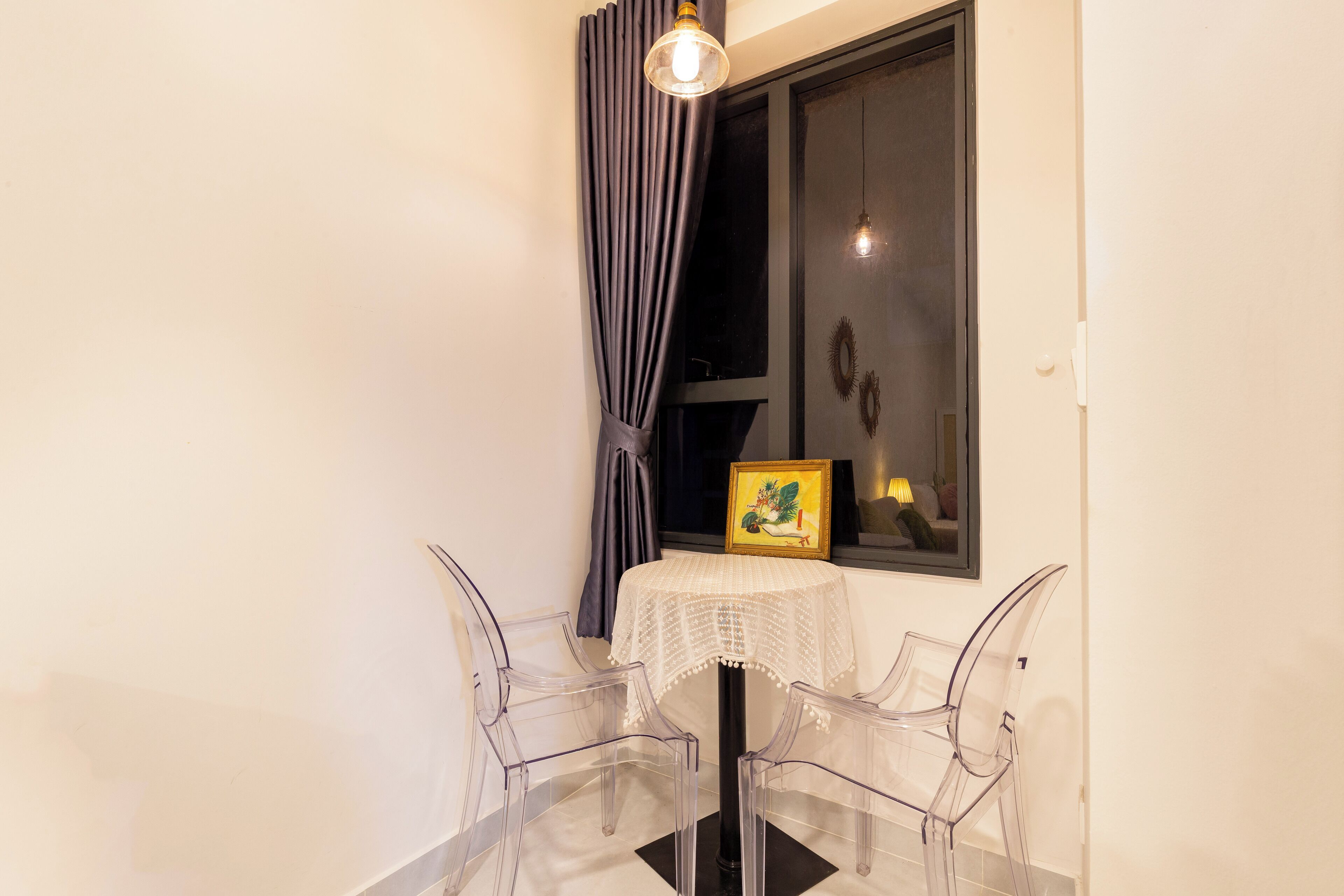 A corner room with a small table and two clear chairs, a window with curtains, and a framed picture on the wall.