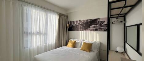 Una habitación de hotel moderna con una cama grande, un escritorio y una silla. Se ve una ventana con cortinas y un televisor montado en la pared.