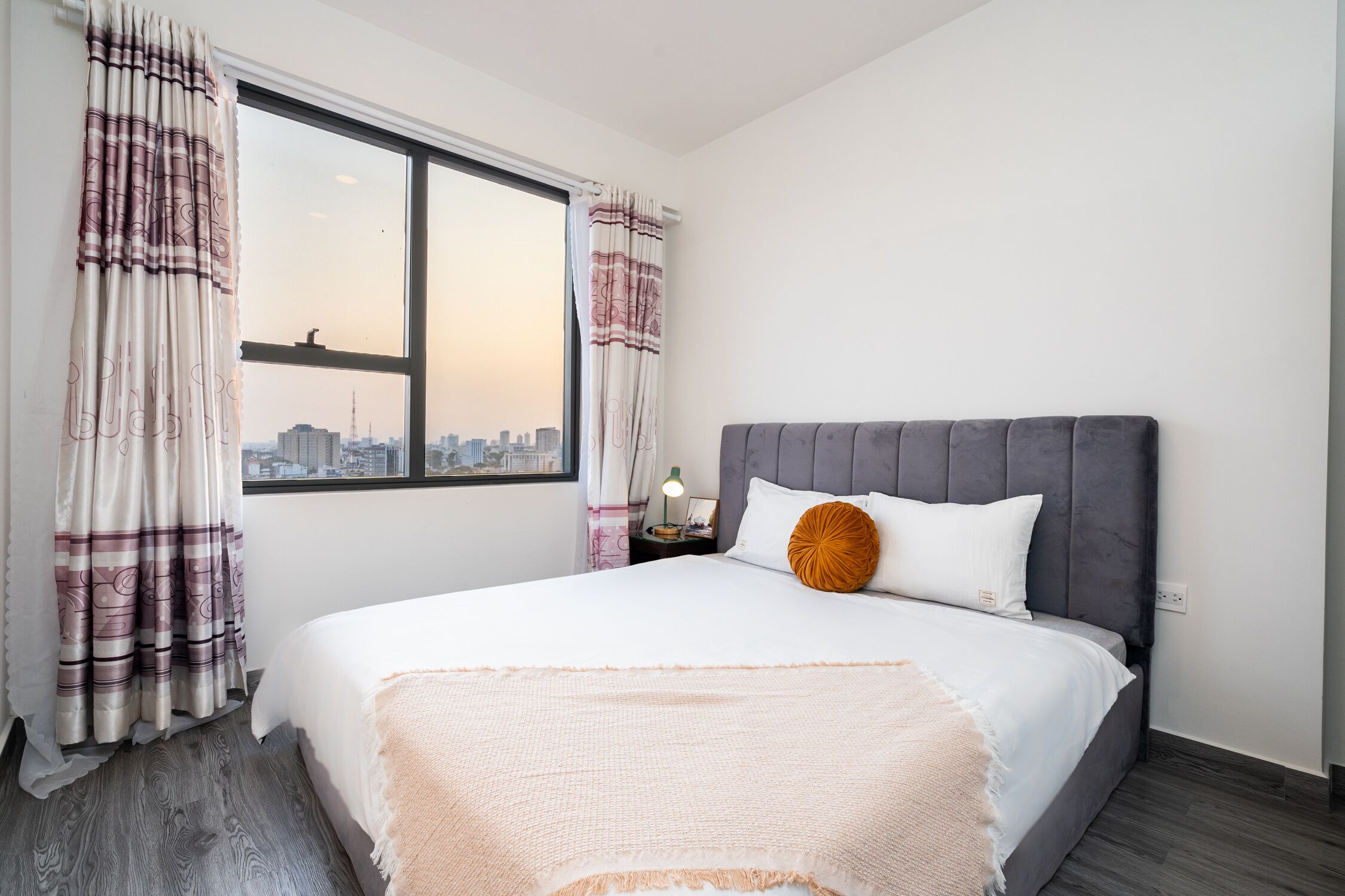 A bedroom with a large window, a bed with a grey headboard, and a city view.