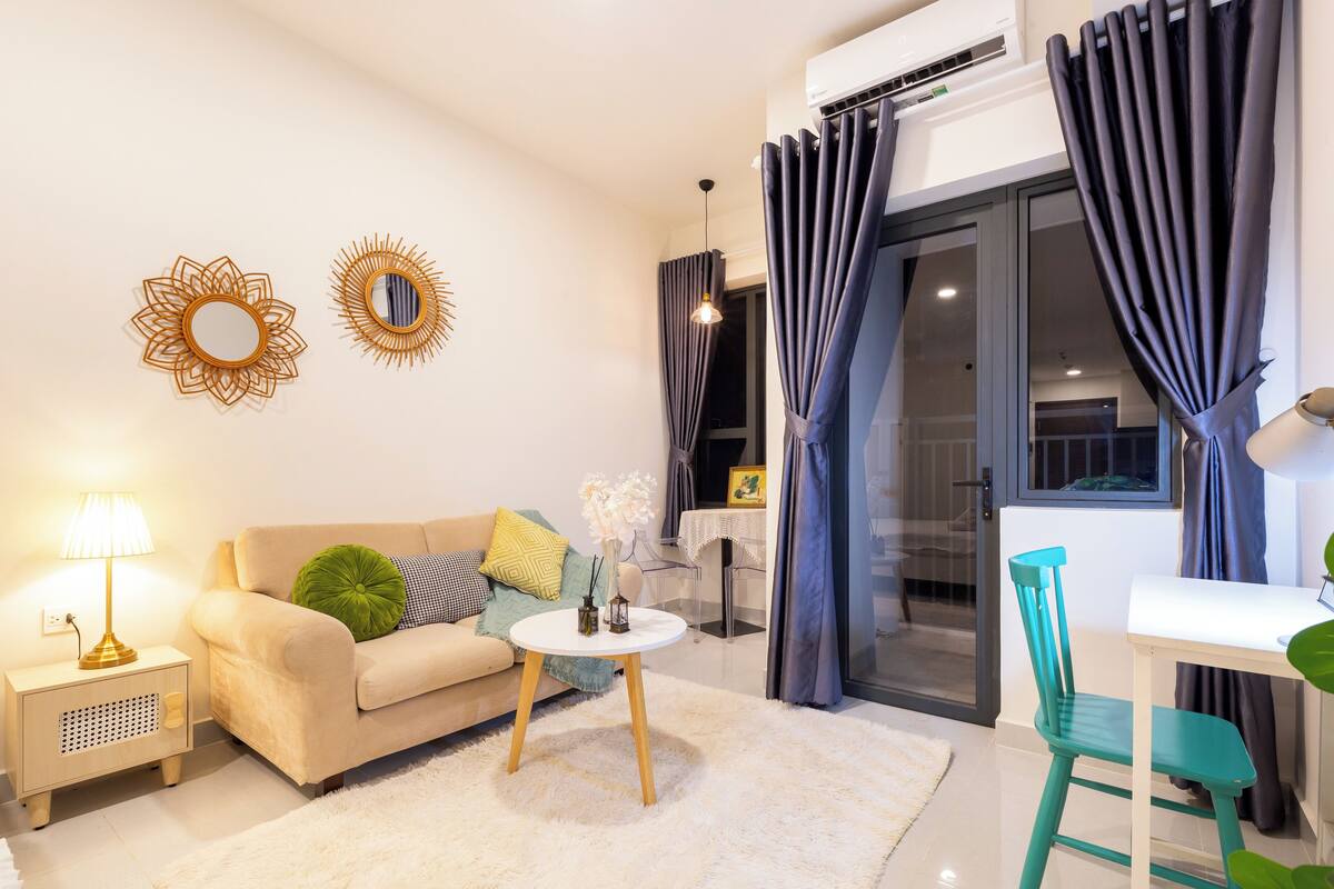 A modern living room with a sofa, a small round table, and a chair. There is a lamp, a mirror, and a window with curtains.