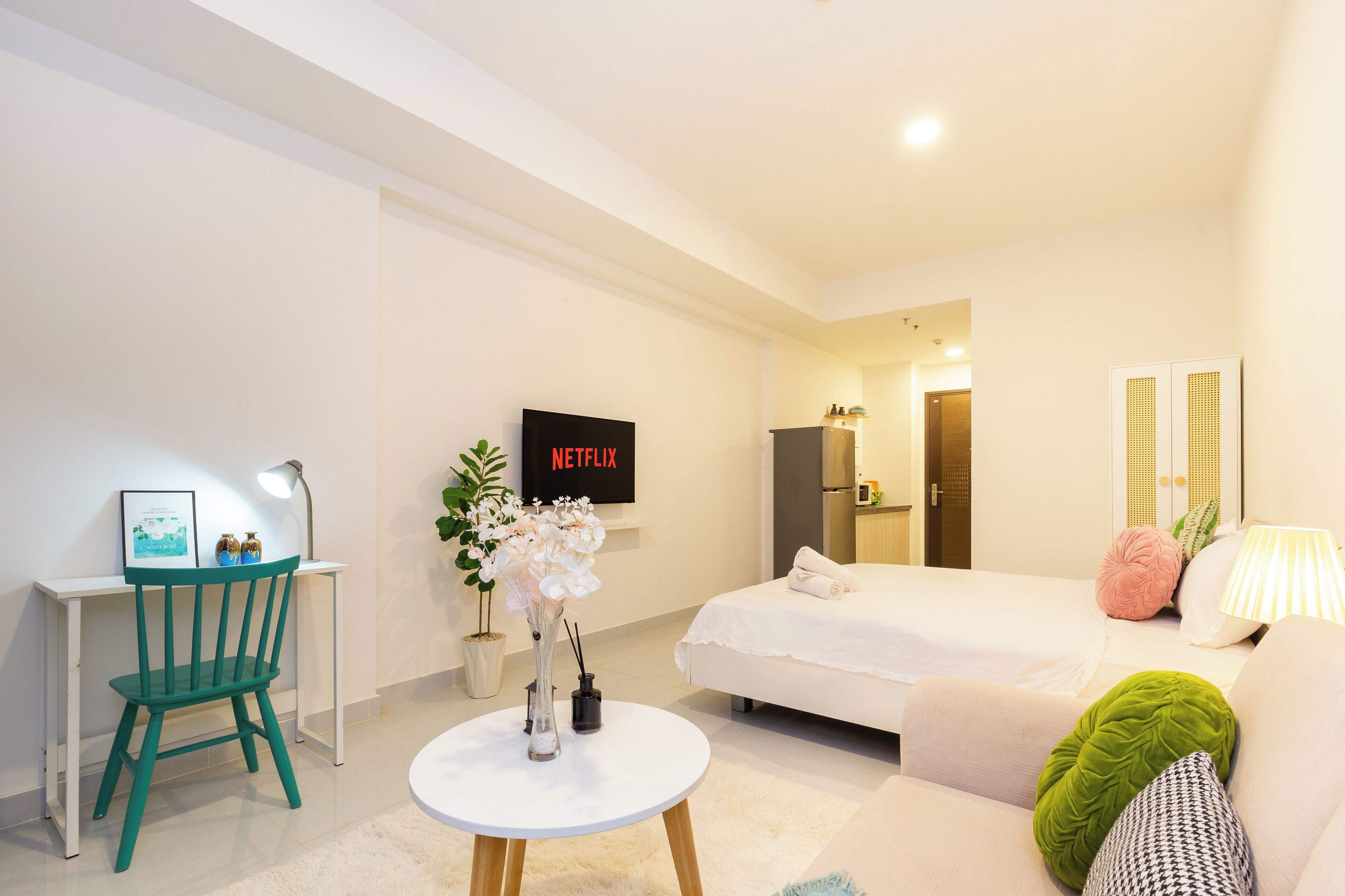 A modern hotel room with a bed, a sofa, a desk with a chair, a TV, and a small table with flowers.
