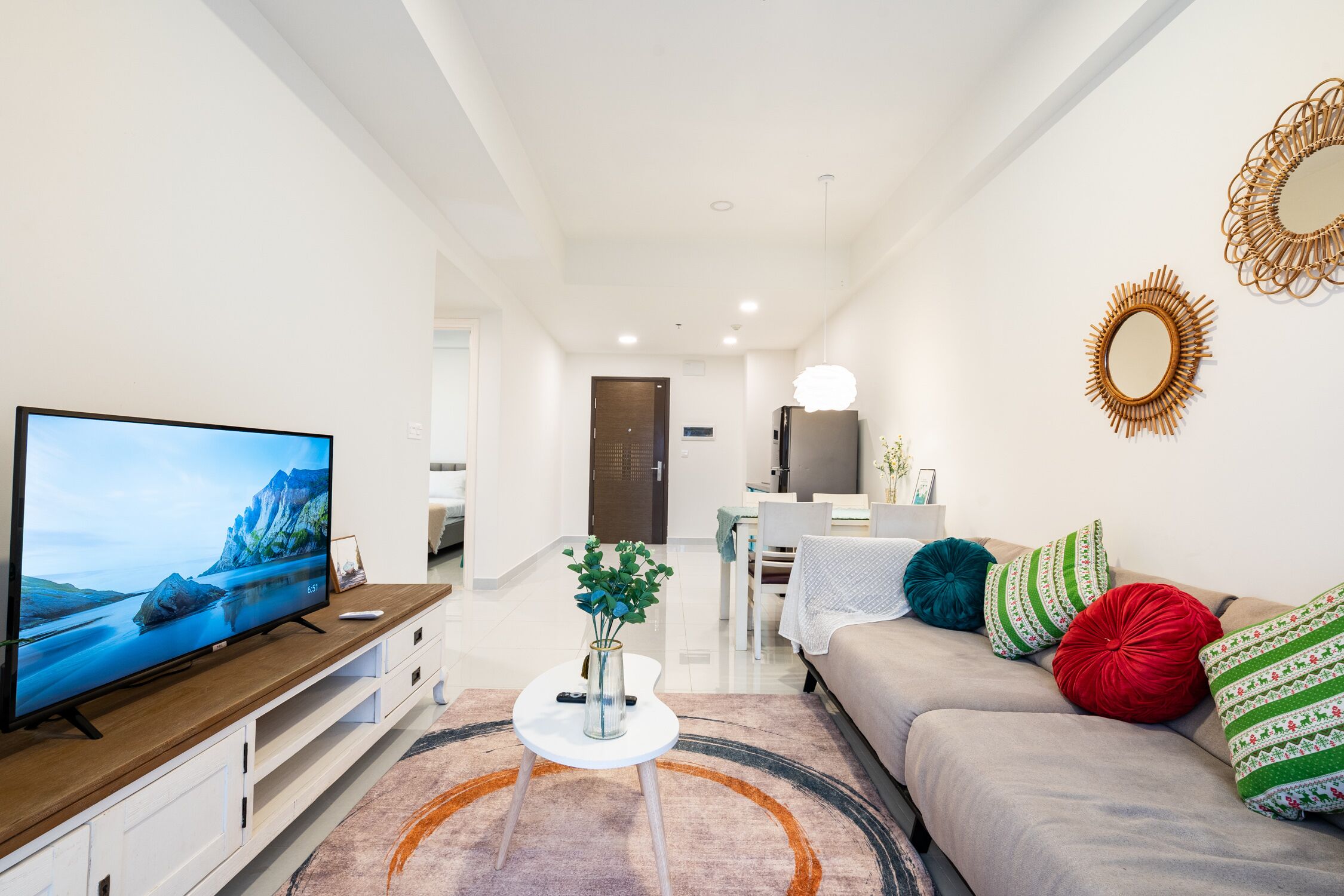 En moderne stue med et fladskærms-TV, et sofabord med en vase, en sofa med farverige puder, og et spiseområde i baggrunden.