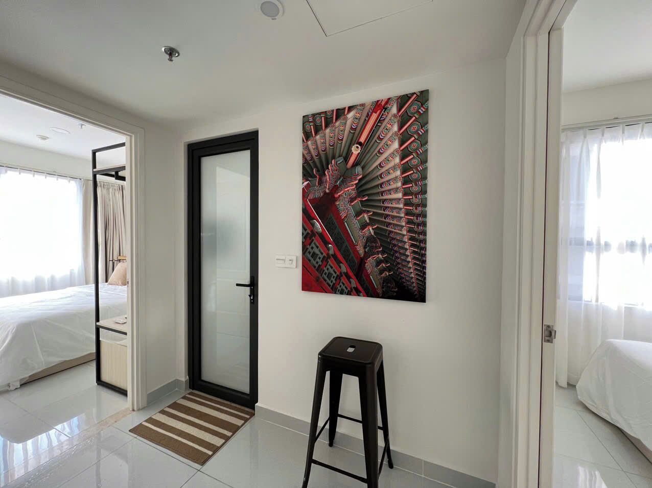 Quarto de hotel moderno com uma cama, uma porta que dá para outro quarto, um banquinho pequeno e um quadro abstrato grande na parede.