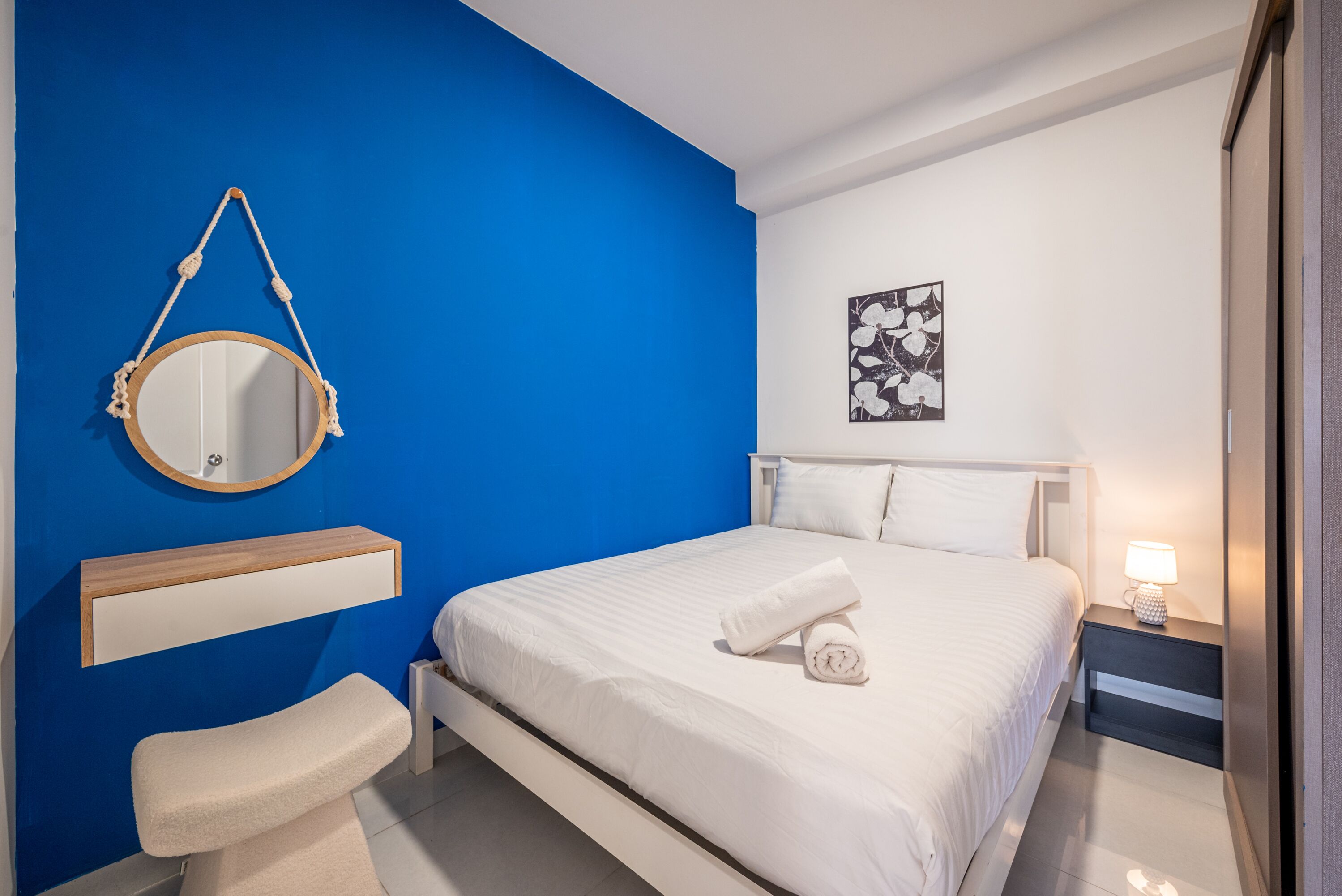 A bedroom with a bed, a bedside table with a lamp, a hanging mirror, and a wall-mounted shelf.