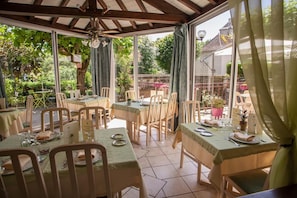 A covered outdoor dining area with tables and chairs, large windows, and a view of the garden. - Logis Auberge de L'ile (Jayac)
