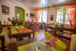 A dining area with multiple tables and chairs, a television mounted on the wall, and a window with curtains. - Logis Auberge de L'ile (Jayac)