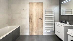 Ein modernes Badezimmer mit Holztür, einer weißen Badewanne, einem weißen Waschtisch mit Spiegel und einem Handtuchhalter.