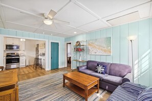 A living room with a purple sofa, a wooden coffee table, and a kitchen area with a microwave and stove.