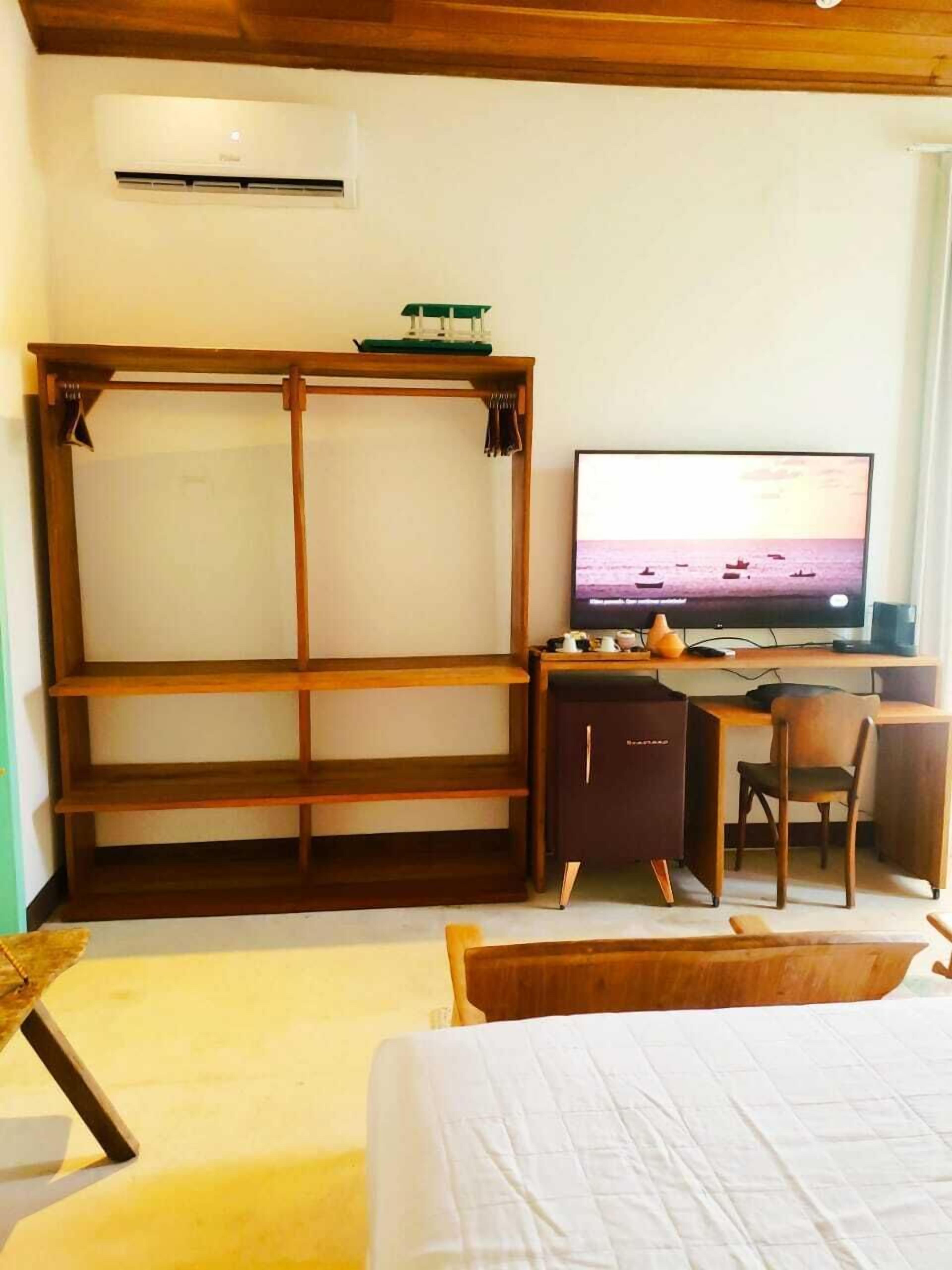 Ein Hotelzimmer mit einem hölzernen Bett, einem Schreibtisch mit Computer, einem Fernseher der eine Strandansicht zeigt und einem Kleiderschrank.