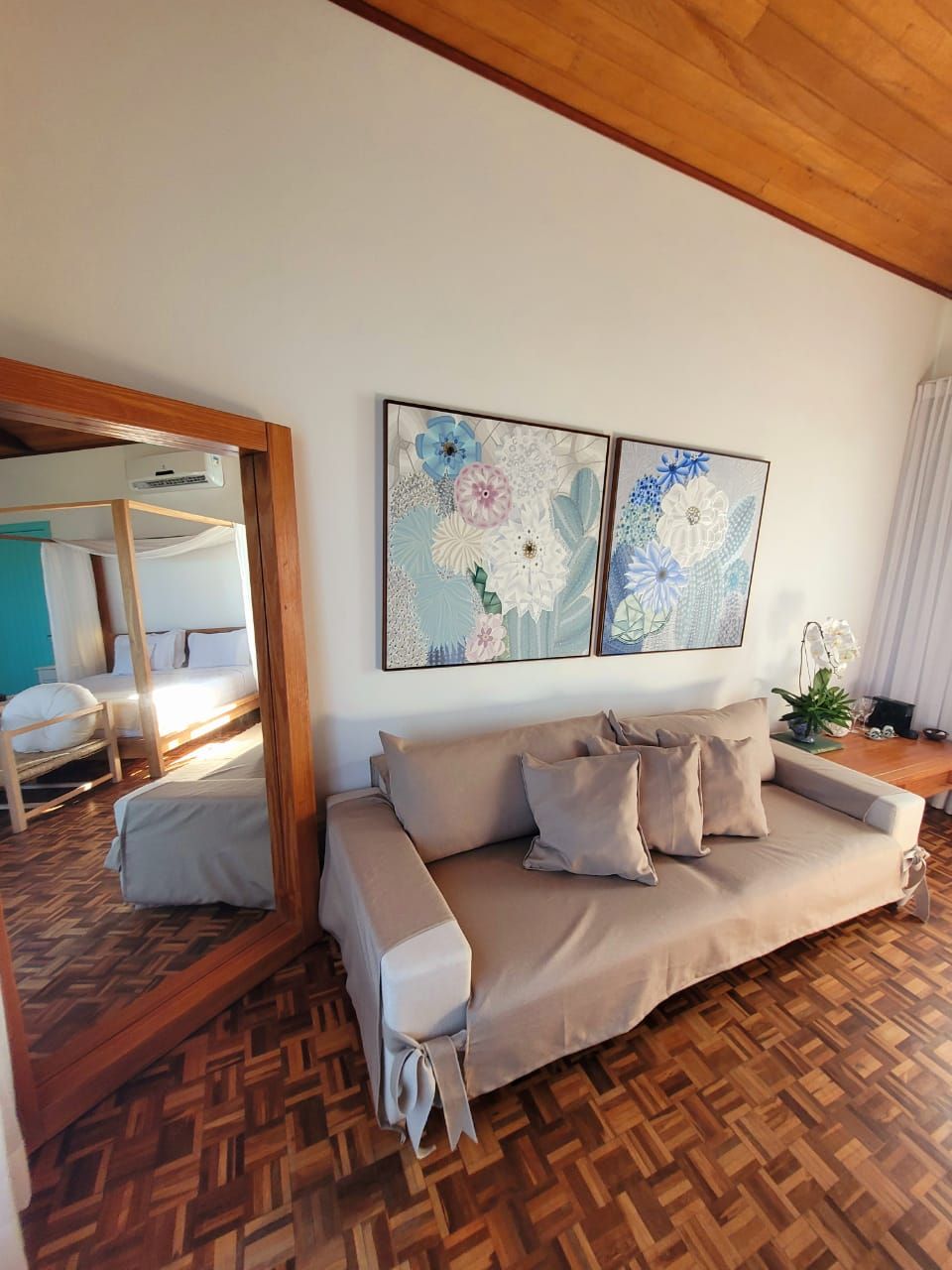 A room with a wooden ceiling, a beige sofa, a large mirror, and two wall-mounted floral paintings.