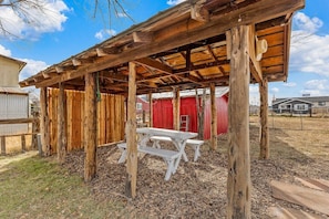 A rustic wooden structure with a picnic table, surrounded by wooden fences and a red building in the background.