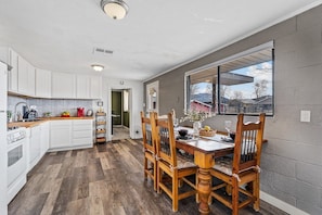 A kitchen with white cabinets, a wooden dining table with chairs, and a window with a view of houses.