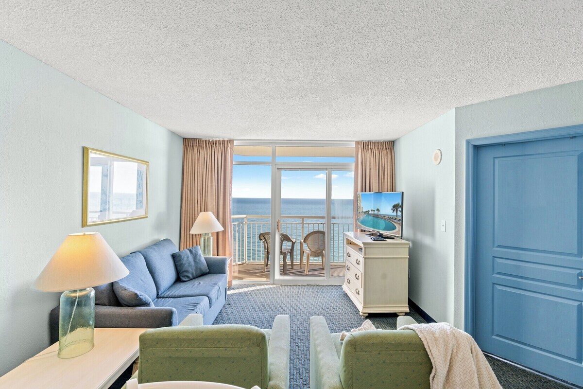 Quarto de hotel com varanda, sofá, televisão e secretária com candeeiro.