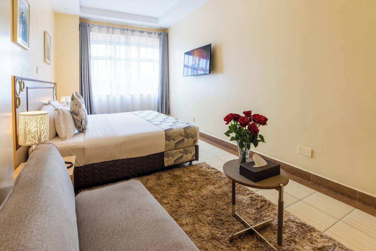 A hotel room with a bed, a sofa, a small table with a vase of flowers, and a television on the wall.