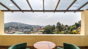 Un balcón con una mesa de madera y dos sillas verdes, con vista a un paisaje pintoresco de un pueblo con edificios y vegetación.