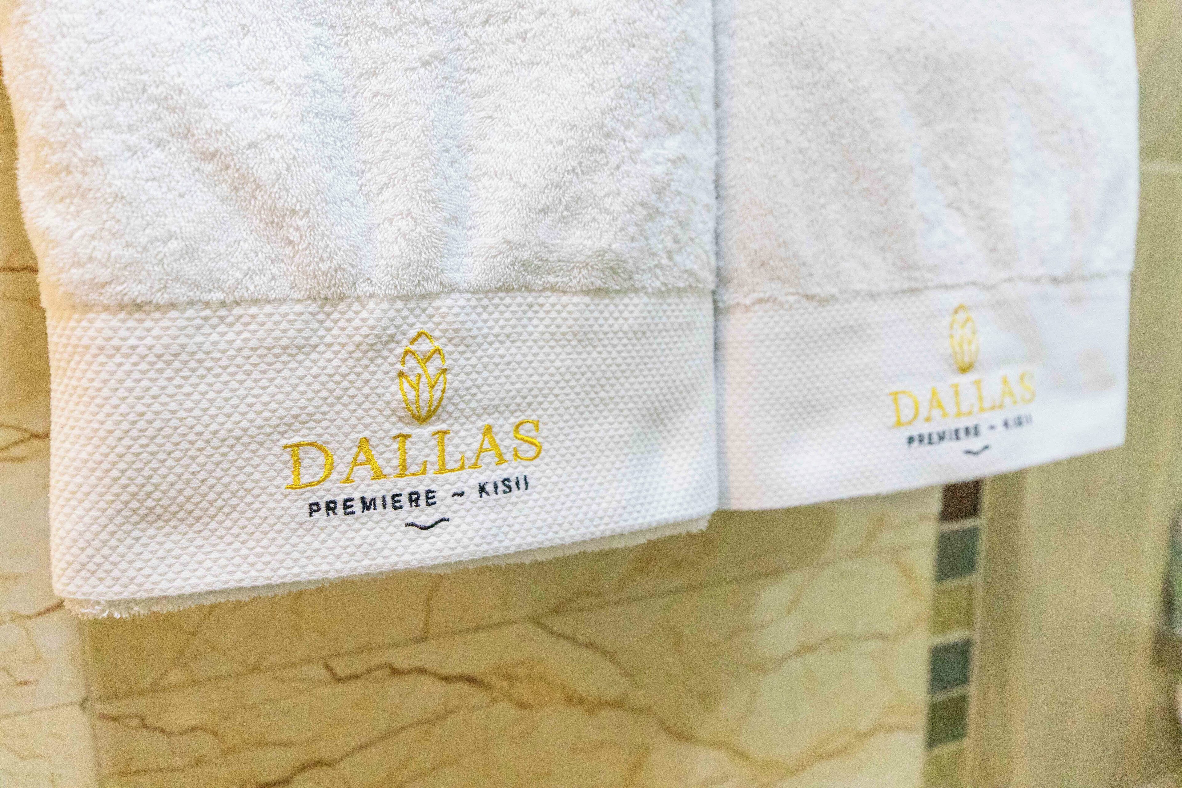 Two white towels with 'DALLAS PREMIERE - KISSI' embroidered on them, hanging on a textured wall.