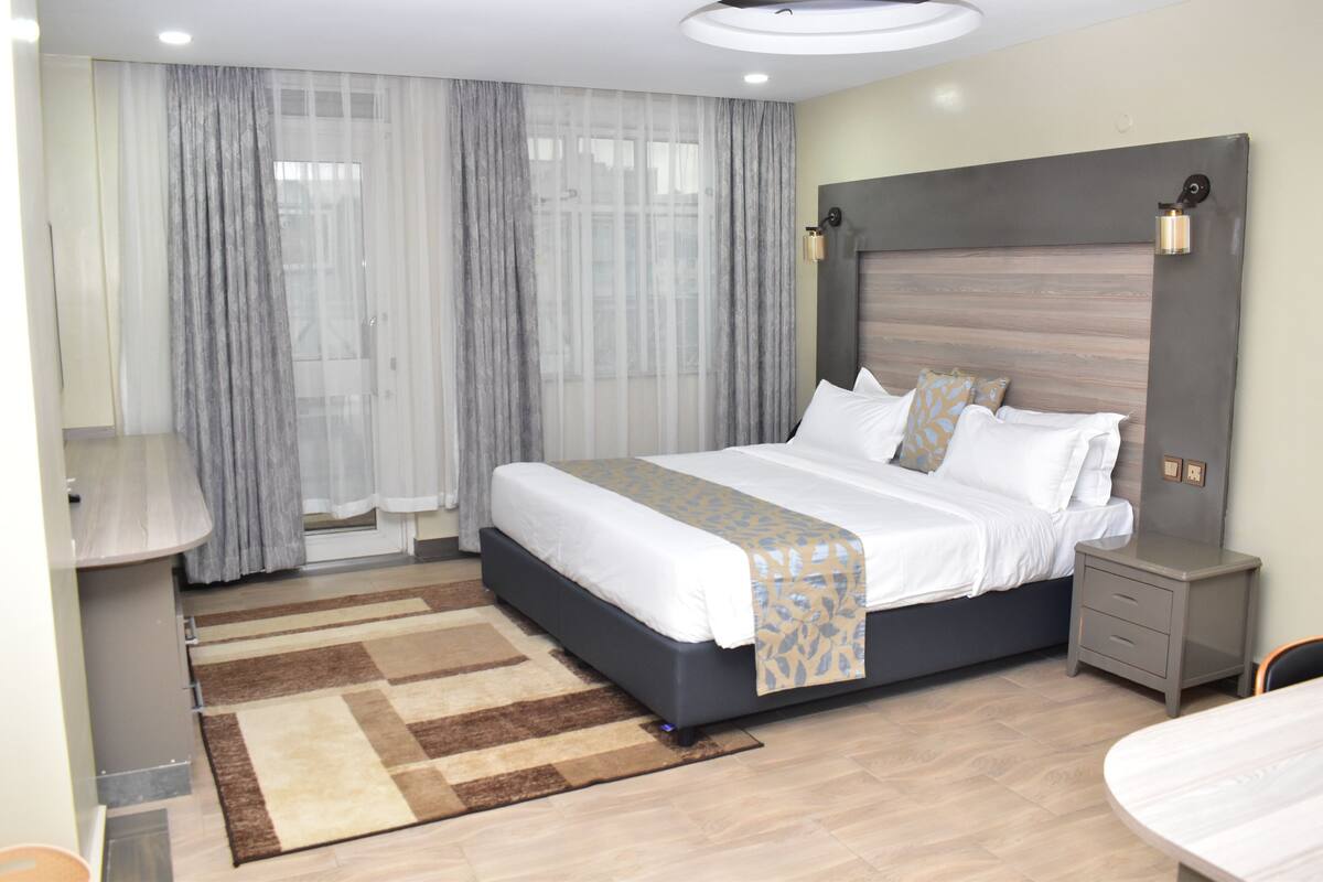 A modern hotel room with a large bed, a bedside table, a desk, and a chair.