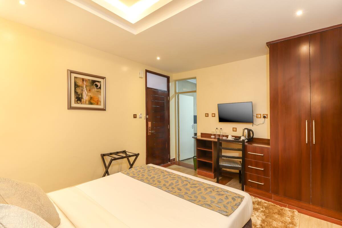 A bedroom with a bed, a wooden wardrobe, a desk with a chair, a television, and a door leading to another room.