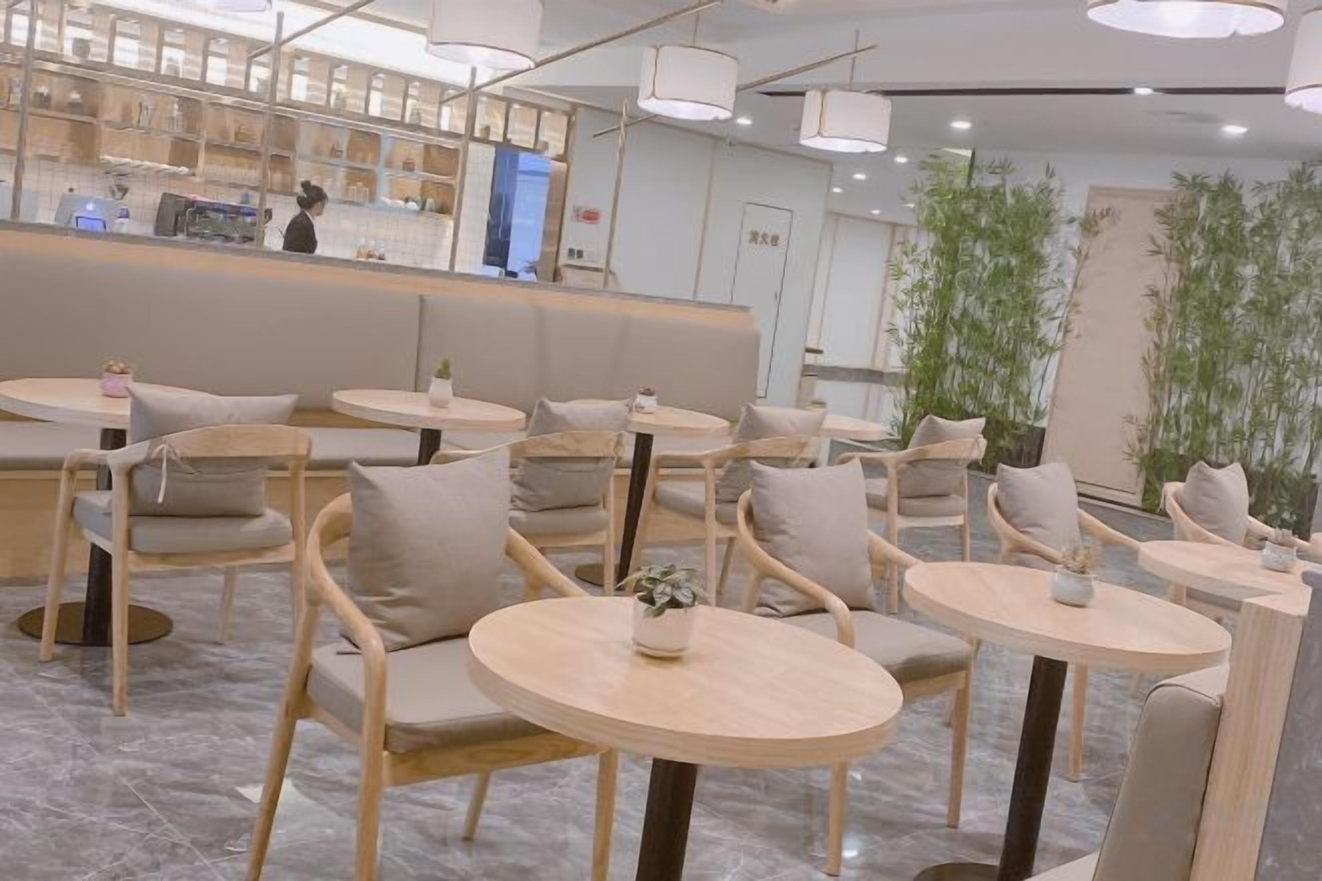 A cafe interior with round tables and cushioned chairs, a counter with a person behind it, and indoor plants.