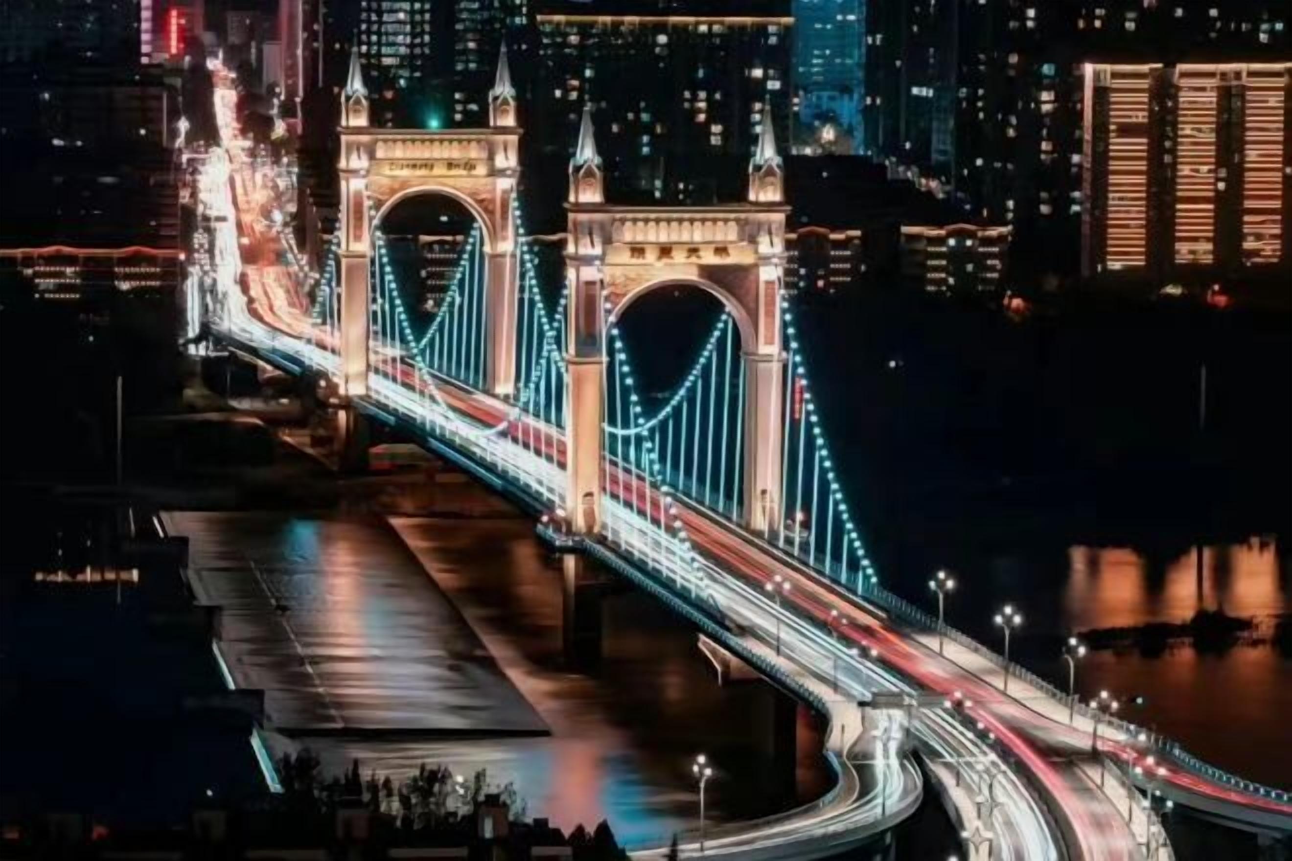 Ein Nachtblick auf eine Brücke mit beleuchteten Kabeln und einer Skyline im Hintergrund.