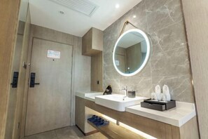 A modern bathroom with a wooden door, a marble wall, a round mirror with a light, a sink with toiletries, and a towel rack.