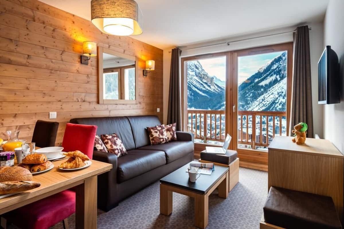 Ein gemütliches Zimmer mit Holzwand, einer Couch, einem Esstisch mit Frühstück und einem großen Fenster mit Bergblick.