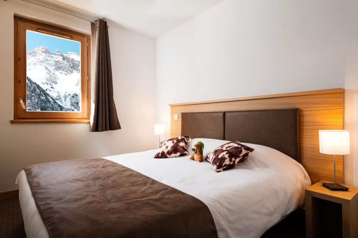 Ein Hotelzimmer mit Bett, Fenster mit Bergblick, Nachttischlampe und Teddybär auf dem Bett.