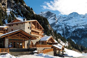 Un chalet de montagne à plusieurs étages, construit en pierre et en bois, doté de balcons et offrant une vue sur les montagnes enneigées.