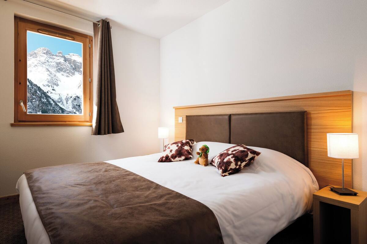 Ein Hotelzimmer mit Bett, einem Fenster mit Vorhängen, einem Nachttisch mit Lampe und einem Teddybären auf dem Bett.