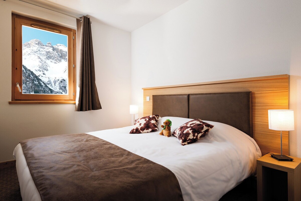 A hotel room with a bed, a window with curtains, a nightstand with a lamp, and a teddy bear on the bed.