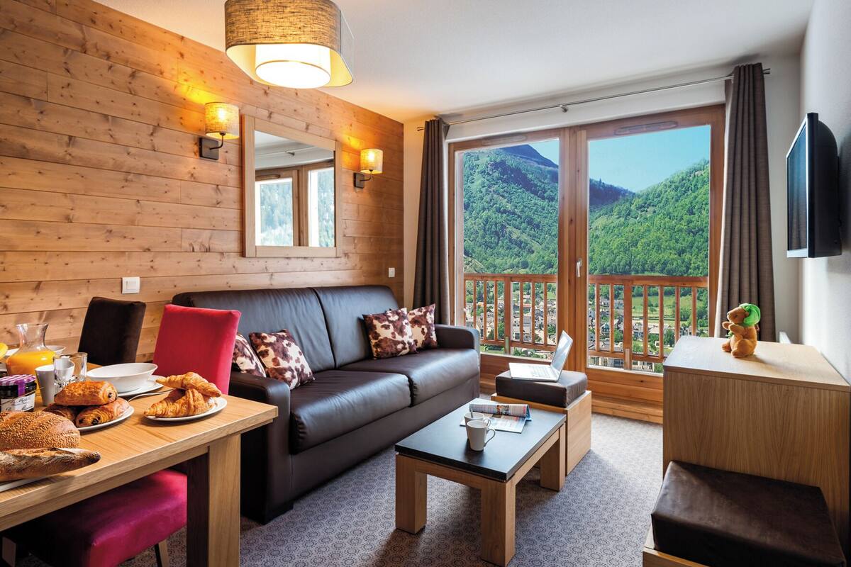 Ein gemütliches Zimmer mit Ledersofa, Esstisch mit Frühstück, einem Laptop und einem Balkon mit Bergblick.