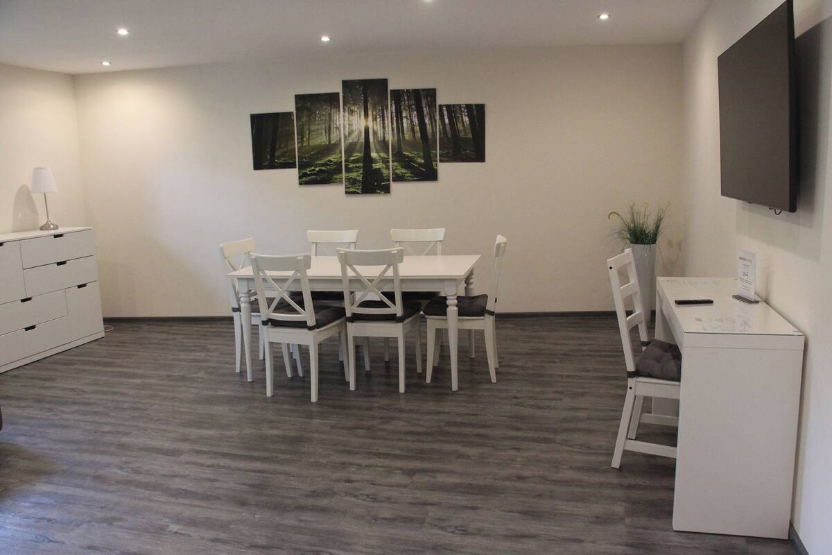 A modern room with a dining table and chairs, a desk with a chair, a flat-screen TV mounted on the wall, and a large forest-themed artwork above the table.