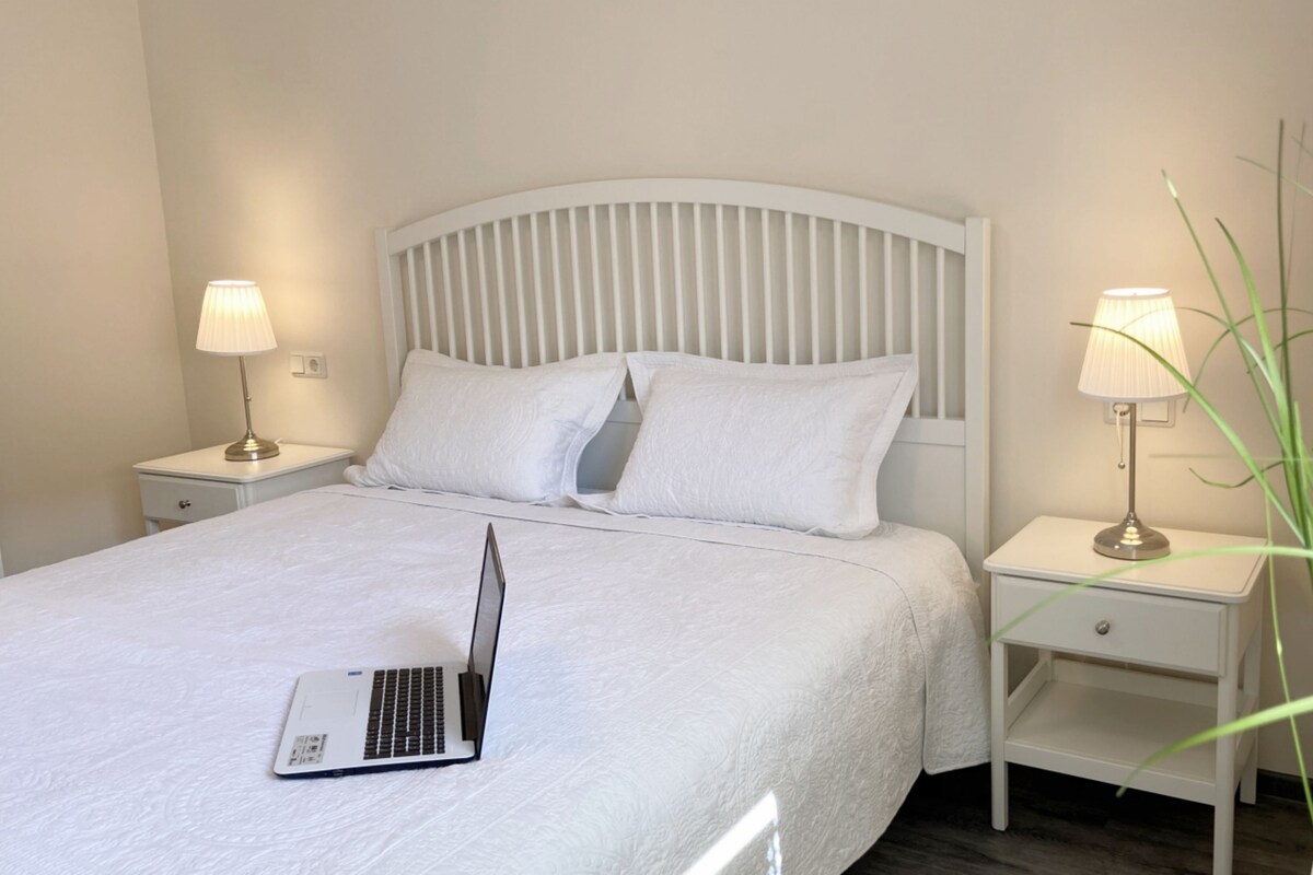 Una cama bien tendida con ropa blanca, un ordenador portátil sobre la cama y dos mesitas de noche con lámparas.