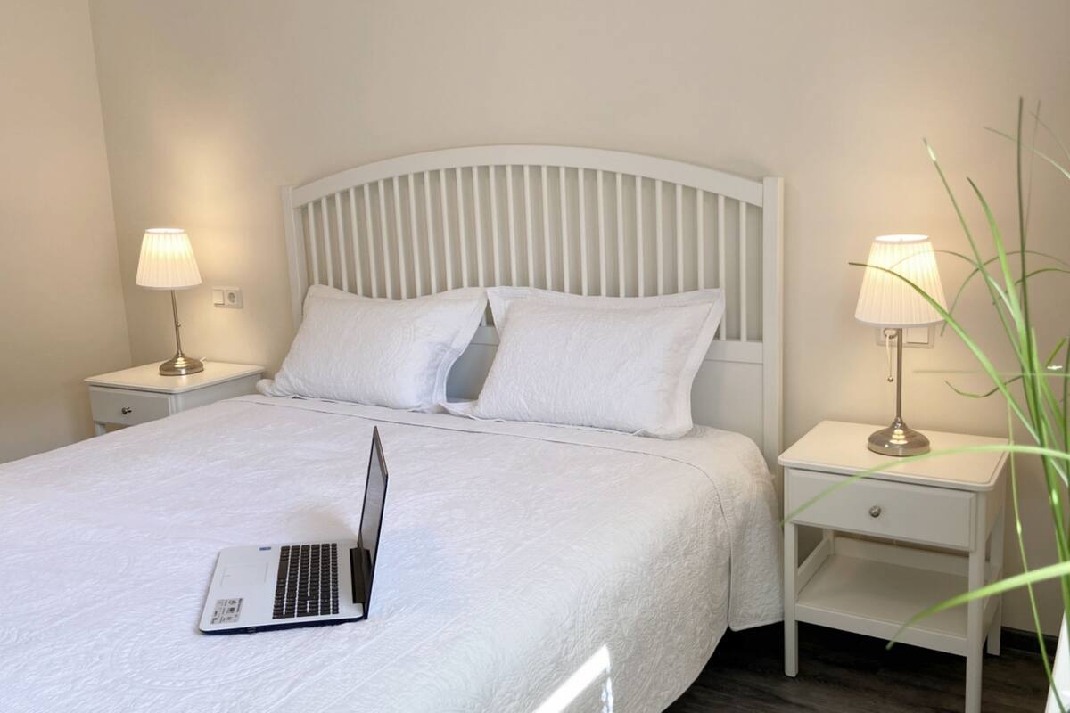 A neatly made bed with white bedding, a laptop on the bed, and two bedside tables with lamps.