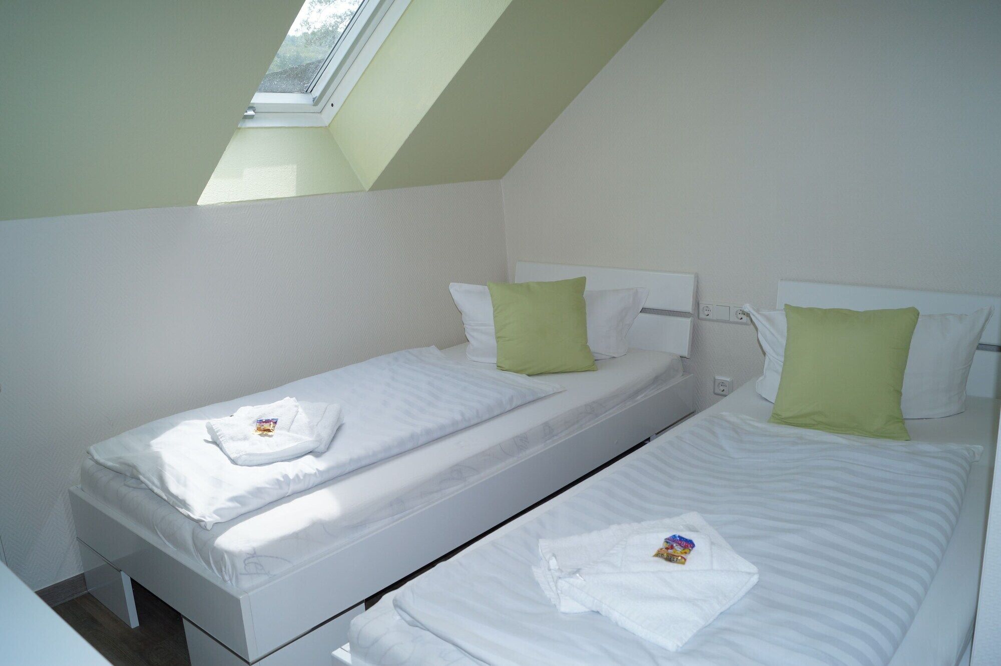 A small, clean room with two single beds, each with white linens and green pillows. A skylight provides natural light.