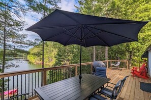 Une terrasse en bois avec une table et des chaises, un grand parasol et une vue sur un lac entouré d’arbres.