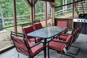 Une terrasse extérieure avec une table en verre et des chaises rembourrées de couleur rouge, un barbecue et une vue sur la forêt.