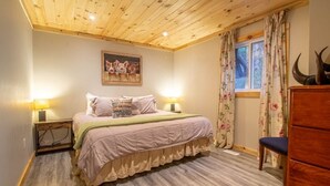 Une chambre avec un plafond en bois, un lit recouvert d’une couvre-lit à motifs floraux, deux lampes de chevet, une fenêtre ornée de rideaux à motifs floraux et une commode en bois.